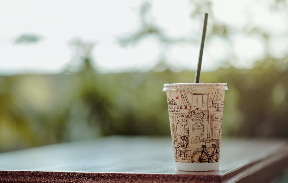 This image shows a strawberry and creme frappuccino with a plastic single-use cup and straw on a busy city street with buildings in the background. The image represents the environmental impact of producing and consuming frappuccinos, particularly the waste generated from disposable cups.